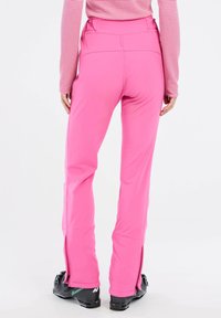 Roze skibroek met een slank model, uitgerust met enkels ritssluitingen en een gladde textuur. Draagt met zwarte skischoenen en een rood gestreept shirt.