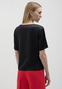 Top corto in raso nero a maniche corte con vestibilità rilassata e scollatura rotonda, indossato con pantaloni larghi rossi. Design semplice senza motivi o decorazioni.