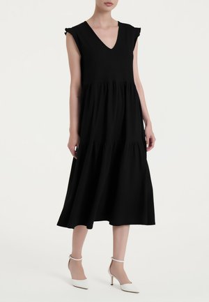 Donna che indossa un vestito midi nero senza maniche con scollo a V e gonna a balze, abbinato a scarpe bianche con tacco alto a punta e cinturini alla caviglia.