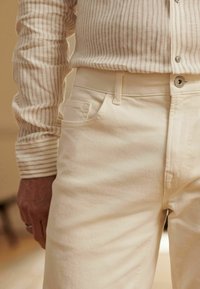 Beige bomuldsbukser med slim-fit design, der har en frontlomme og subtil tekstur; parret med en stribet, lys skjorte.