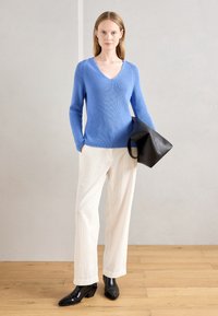 Maglione blu a costine con scollo a V, abbinato a pantaloni larghi in corduroy bianchi e stivali neri a punta con caviglia. Borsa nera in mano.