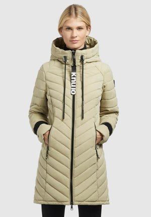Manteau long matelassé beige avec capuche, fermeture éclair sur le devant et cordons de serrage. Présente des accents noirs et deux poches latérales. Texture légèrement brillante.