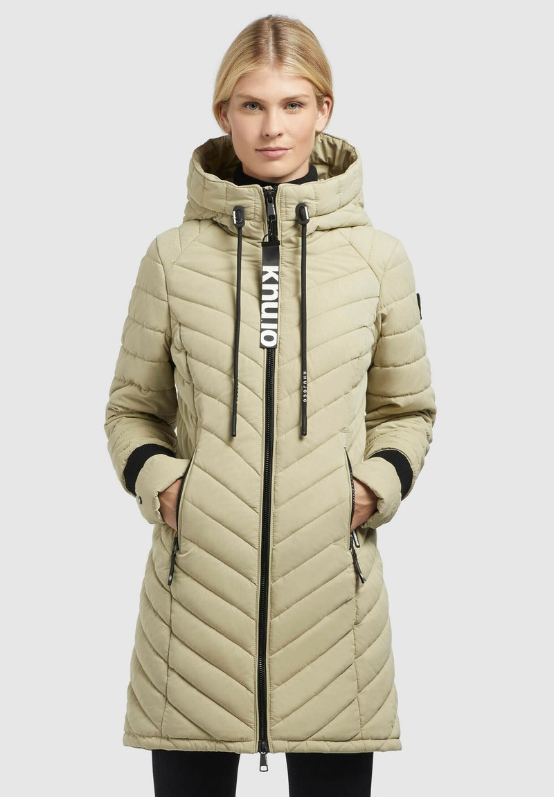Beige gesteppter langer Mantel mit Kapuze, Frontreißverschluss und Kordelzügen. Verfügt über schwarze Akzente und zwei seitliche Taschen. Leichte schimmernde Textur.