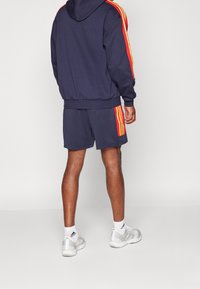 Námořnická mikina s červenými a žlutými pruhy podél ramen, spárovaná s odpovídajícími šortkami. Materiál vypadá měkce; zahrnuje sportovní boty a ponožky.