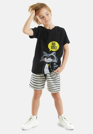 Schwarzes T-Shirt mit einem Waschbär-Graphic und einer Sprechblase "Hi BRO". Kombiniert mit gestreiften grauen Shorts und weißen High-Top-Sneakern.