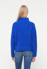 Un pull en laine bleu avec un col montant, un motif texturé et des manches tombantes, associé à un jean en denim bleu.