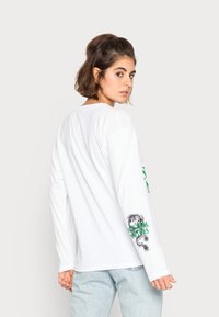 T-shirt branca de mangas longas com decote redondo; apresenta texto verde e um gráfico de dragão preto na manga esquerda, combinado com jeans de denim claros.