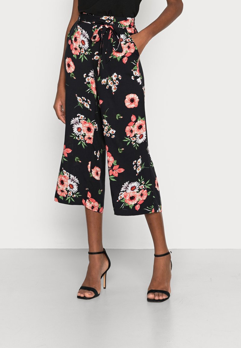 Pantalon large noir raccourci avec imprimé floral rose et blanc, taille nouée, porté avec des sandales à talons noires et un haut noir.