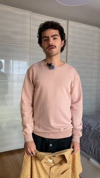 Jeune homme aux cheveux bouclés et moustachu, portant un sweat-shirt rose, tient une chemise jaune à carreaux à l'intérieur près de portes coulissantes blanches.