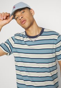 La maglietta a righe presenta righe blu, verdi, bianche e rosa, con un colletto blu navy. È accessorizaria con un cappellino azzurro e una collana in argento.