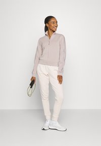 Light purple zip-up jacket with ribbed cuffs, paired with cream jogger pants. Model holds a jump rope, wearing white athletic shoes.