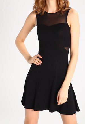 Woman wearing a sleeveless black dress with sheer mesh panels at the neckline and waist, standing with one hand on hip.