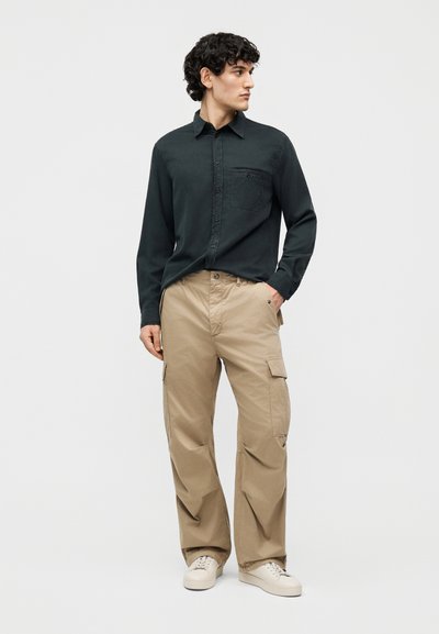 Άνδρας που φοράει σκούρο πράσινο πουκάμισο με κουμπιά, μπεζ παντελόνι καμπάνιας και λευκά αθλητικά παπούτσια, στέκεται με το ένα χέρι στην τσέπη, κοιτάζοντας προς τα πλάγια.