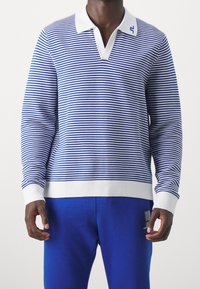 Pitkähihaista raidallista polo-takkia sinivalkoisena. Varustettu valkoisella kauluksella ja ribatulla hihansuulla. Yhdistetty solidinsinisiin housuihin.