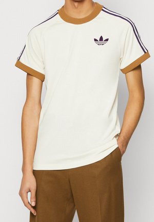 Man draagt een wit T-shirt met korte mouwen met een bruine kraag en manchetten, paarse strepen op de schouders en een bruine broek, hand in de zak.