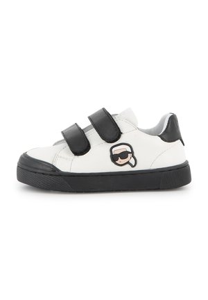 Baskets blanches pour enfants avec semelle noire et deux bandes velcro noires, avec un petit visage de dessin animé avec des lunettes de soleil sur le côté.