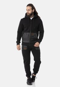 Schwarze Kapuzenjacke mit einer glatten, satinähnlichen Tasche und kontrastierenden Stoffdetails, kombiniert mit schwarzen Jogginghosen, die ähnliche Akzente aufweisen.