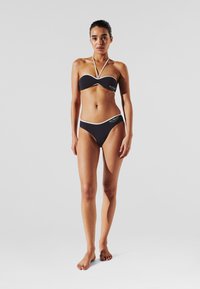 Crni bikini s bijelim detaljima. Gornji dio ima halterski vrat i potporu. Donji dio ima nisku liniju. Glatka tekstura, minimalističko brendiranje.