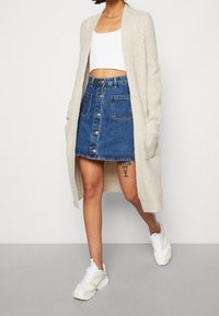 Leichtes beige gestricktes Cardigan, weißes Cropped-Top, Denim A-Linien-Rock mit Knöpfen und Taschen, weiße Sneaker. Geschmeidige Textur und lässige Passform.