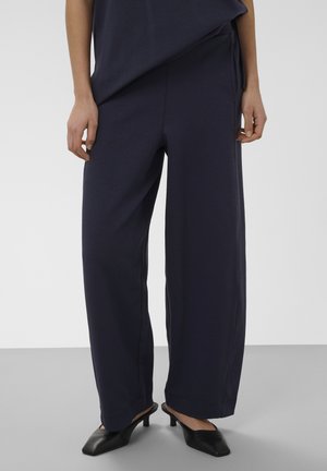 Femme vêtue d'un pantalon ample bleu marine et de chaussures noires à talons bas et bouts pointus, debout devant un fond gris et blanc uni.