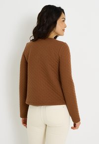 Chaqueta marrón acolchada con mangas largas, que presenta un escote redondeado y un patrón texturizado de diamantes. Llevada sobre jeans de color claro.