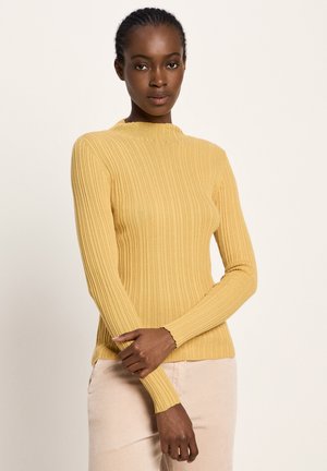 Frau mit geflochtenem Haar, die einen langen senfgelben gerippten Pullover und eine hellbeige Hose trägt, steht vor einem schlichten hellen Hintergrund.