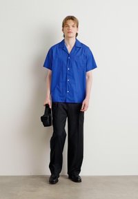 Camisa azul de botão com mangas curtas e bolso, combinada com calças pretas e sapatos pretos, segurando uma câmara preta. Fundo liso.