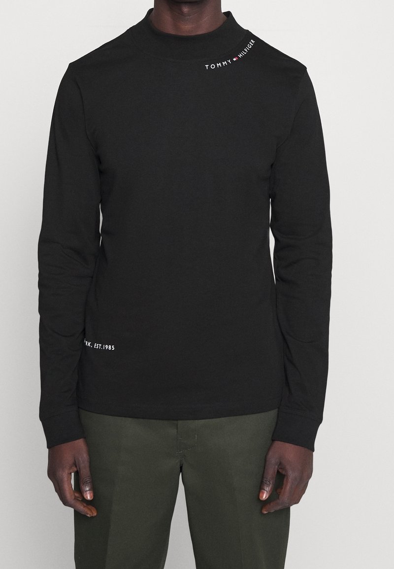 Svart långärmad skjorta med "Tommy Hilfiger"-logotyp på högra axeln och gröna byxor bärs av en stående person.