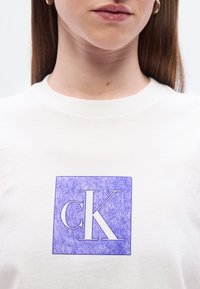 Λευκό βαμβακερό t-shirt με ένα ανοιχτό μωβ τετράγωνο που περιέχει τα γράμ ματα "cK" σε άσπρο, παρουσιάζοντας ένα υφασμάτινο σχέδιο μέσα στο τετράγωνο.