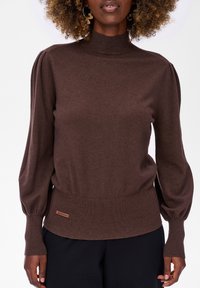 Ciemnobrązowy długi sweter dzianinowy z wysokim kołnierzem i bufiastymi rękawami noszony przez osobę z kręconymi włosami na białym tle.