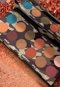 Palette im Camouflage-Muster mit 12 Lidschattenfarben in verschiedenen Texturen. Die Farben umfassen Teal, Gold, Braun, Grün und Pink, mit matten und schimmernden Finishes.