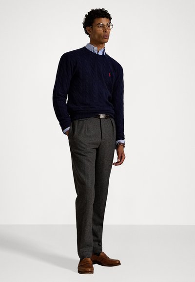 Marinefarbener Kabelstrickpullover, graue plissierte Hose, braune Lederschuhe, blau-weiße kariertes Hemd, das am Kragen hervorschaut, und eine runde Brille.