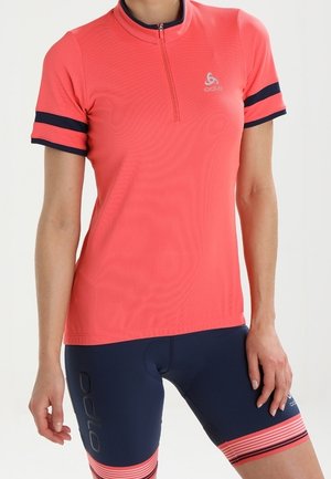 Kvinde iført en koral kortærmet cykeltrøje med halv lynlås og marineblå cykelshorts med koralstribedetaljer.