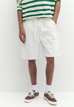 Mężczyzna w białych szortach typu cargo, zielono-białej paskowanej koszulce z długim rękawem, białych skarpetkach i brązowych tenisówkach z materiału stojący.