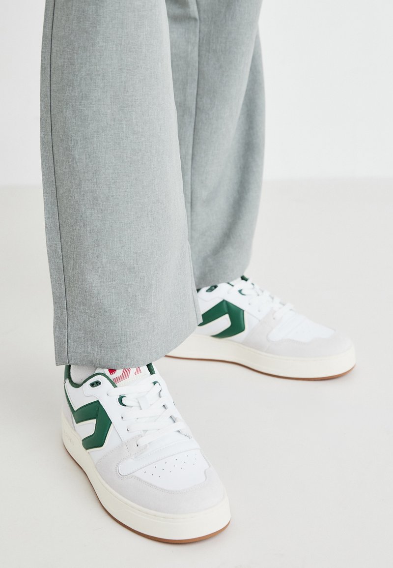 Sneaker bianchi e verdi realizzati in pelle liscia e suede, con un accento del logo verde ben in evidenza e lacci bianchi. Pantaloni grigi parzialmente visibili.