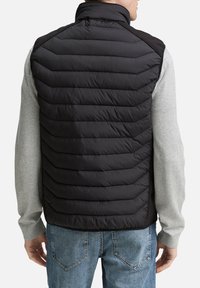 Chaleco acolchado negro con costuras horizontales, cuello alto y material contrastante en los hombros. Se lleva sobre una camiseta de manga larga gris claro.