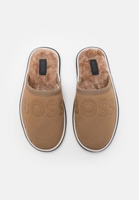 Chaussons beiges avec un intérieur doux et pelucheux. Présentent un logo "BOSS" en relief, un design arrondi et une bordure noire contrastante sur la semelle.