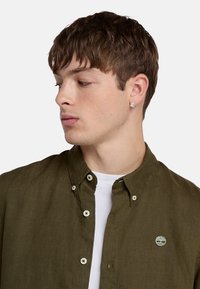 Camisa de lino verde oliva con botones y una camiseta blanca por debajo, con un pequeño logo bordado en el pecho y botones visibles.