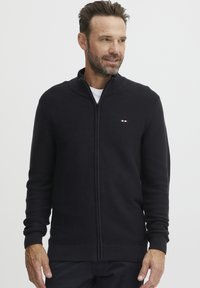 Fekete cipzáras pulóver bordázott textúrával, magas gallérral és elasztikus mandzsettákkal. Kicsi logó részlet piros, fehér és kék színben a mellkason.