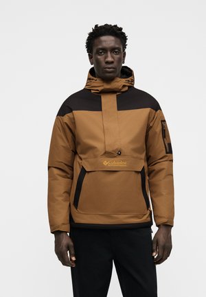Homme portant une veste à capuche Columbia Sportswear marron et noire avec une poche frontale et une poche zippée sur la manche, debout devant un fond uni.