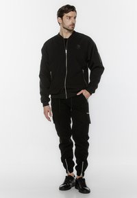 Czarna kurtka bomberka z zapięciem na zamek, ściągaczami przy rękawach i bocznymi kieszeniami, zestawiona z dopasowanymi czarnymi spodniami z zamkami błyskawicznymi przy nogawkach i kieszeniami cargo.