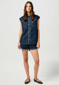 Camicia senza maniche in denim scuro con colletto appuntito, due tasche sul petto e cuciture bianche; abbinata a pantaloni shorts in denim scuro. Ballerine nere.