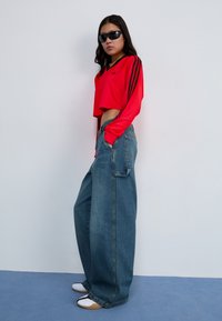 Rotes, kurzes Langarmshirt mit schwarzen Streifen, kombiniert mit weit geschnittenen blauen Jeans. Model trägt schwarze Sonnenbrillen und zweifarbige Schuhe.