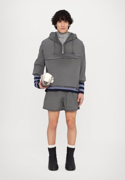 Felpa grigia oversize con collo zippato, abbinata a pantaloni corti grigi. Presenta accenti a strisce blu sulle maniche e stivaletti neri alla caviglia. Sta tenendo una borsa piccola.