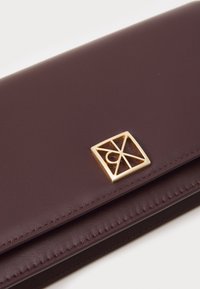 Maroon nahkainen lompakko, jossa on sileä pinta, etupuolella kultainen geometrinen logo ja ommellut reunat hienostuneen viimeistelyn saavuttamiseksi.