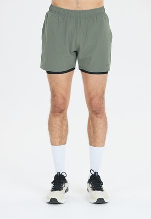 Grønne atletiske shorts med sort kant, der har en elastisk talje. Båret med hvide sokker og sorte og cremefarvede sneakers.