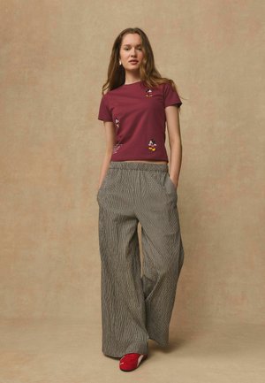 Junge Frau trägt ein bordeauxrotes Mickey Mouse T-Shirt, weit geschnittene karierte Hose, rote Turnschuhe, steht mit den Händen in den Taschen vor einem beigen Hintergrund.