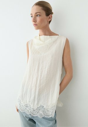 Kvinde med lyst brunt hår samlet bagpå iført hvid ærmeløs top med blonde detaljer ved hals og kant, kombineret med lyseblå jeans.