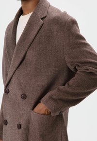 Manteau croisé marron en mélange de laine avec un col cranté, deux poches avant et des boutons marron foncé. Le tissu a une texture douce.