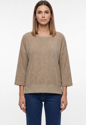 Woman with straight shoulder-length hair wearing a loose beige long-sleeve top and blue jeans, standing against a plain white background.